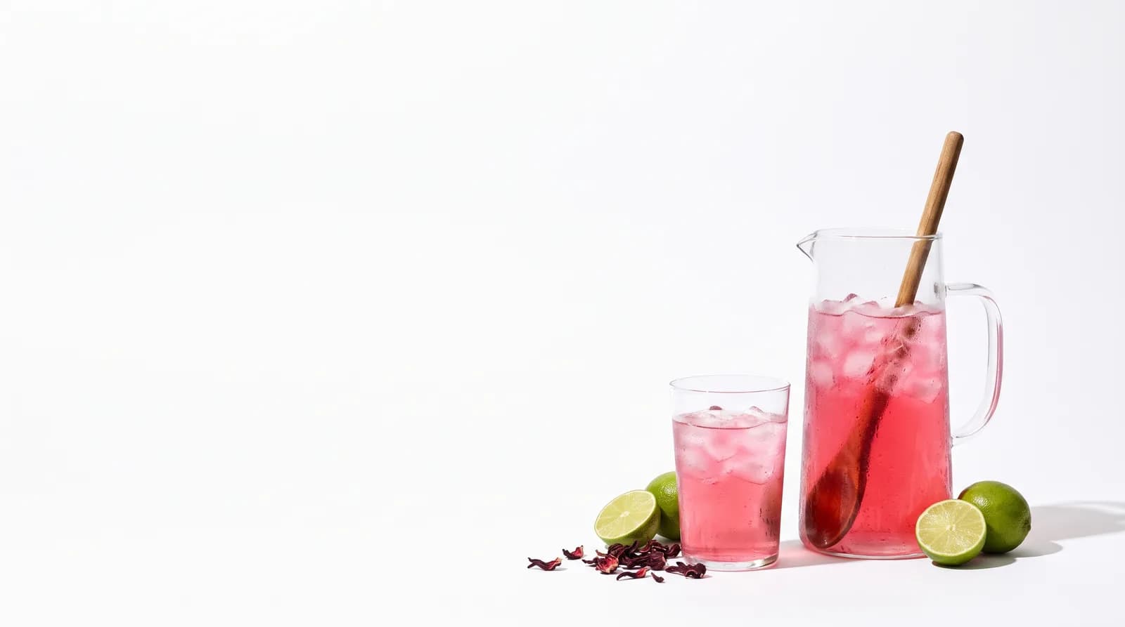 Una jarra de agua de jamaica con hielo y un vaso servido, con limones y flor de jamaica al lado.
